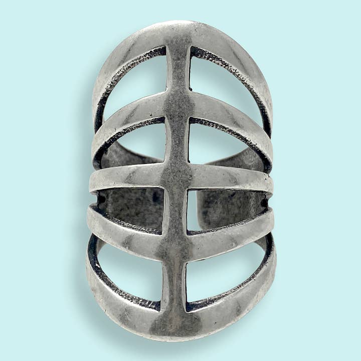 The Silver Gladiator Ring by Ornamental Things features a wide, silver-toned metal band with an openwork design of horizontal and vertical cut-outs, reminiscent of gladiator sandals, elegantly displayed on a light blue background.