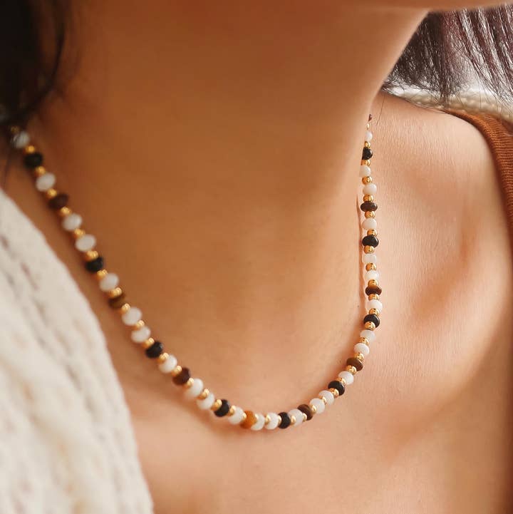 The Arrow Boho Beaded Necklace by Flanerie by MAIQ is a hypoallergenic piece featuring alternating white, black, and translucent brown beads with 18K gold-toned spacers, worn around a person’s neck.