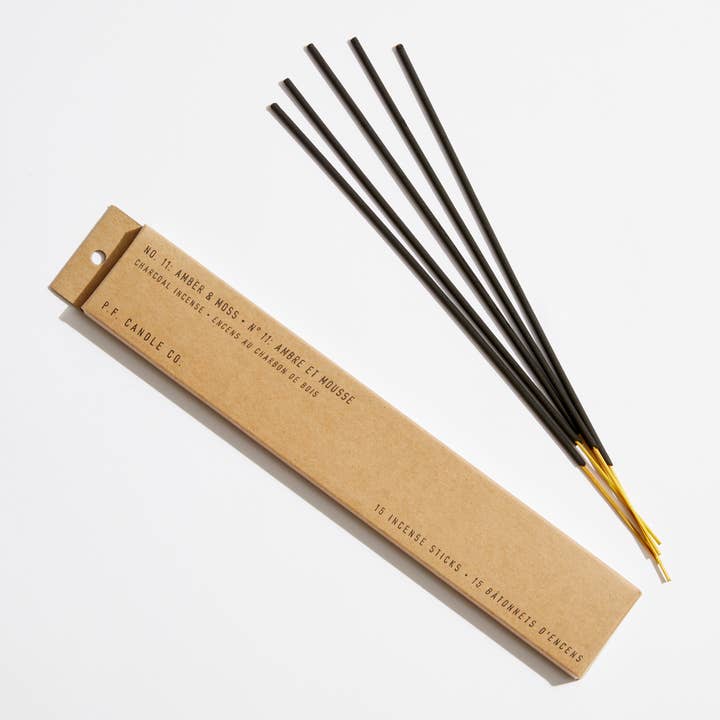 A brown box labeled “Amber & Moss Incense Sticks” by P.F. Candle Co. sits next to four black charcoal incense sticks with gold-colored ends, all arranged on a white background.