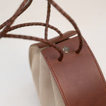Close-up of the Joi Bag by Stash, showcasing Japanese-inspired design with brown leather accents, braided leather handles, beige sand roughout suede sides, and a small metal button detail.