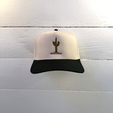 The Shop Hat | Saguaro by Ballad of the Bird Dog, a beige and black cap with an embroidered green cactus and "SAGUARO," is shown against a white wooden wall.