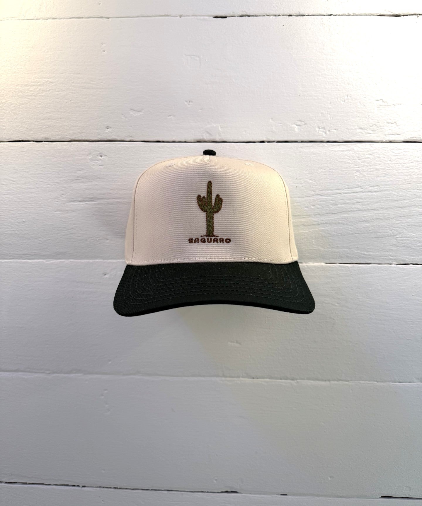 The Shop Hat | Saguaro by Ballad of the Bird Dog, a beige and black cap with an embroidered green cactus and "SAGUARO," is shown against a white wooden wall.