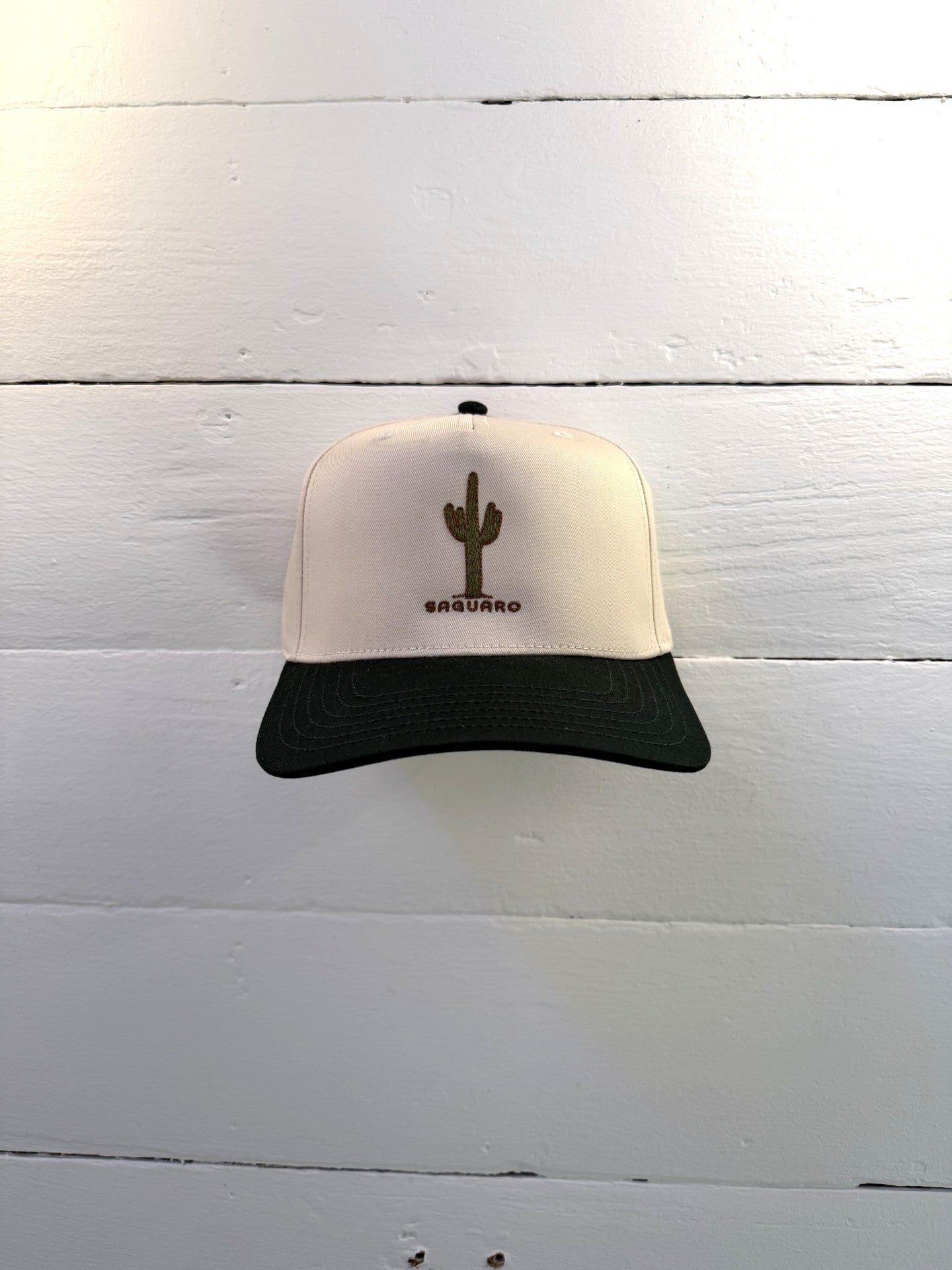 The Shop Hat | Saguaro by Ballad of the Bird Dog, a beige and black cap with an embroidered green cactus and "SAGUARO," is shown against a white wooden wall.