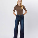 A woman stands against a plain background in a fitted brown top, heeled sandals, and Vervet's 90's Vintage Super High Rise Dark Wash Loose Jeans with wide legs, hands tucked into her pockets.