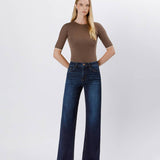 A woman stands against a plain background in a fitted brown top, heeled sandals, and Vervet's 90's Vintage Super High Rise Dark Wash Loose Jeans with wide legs, hands tucked into her pockets.