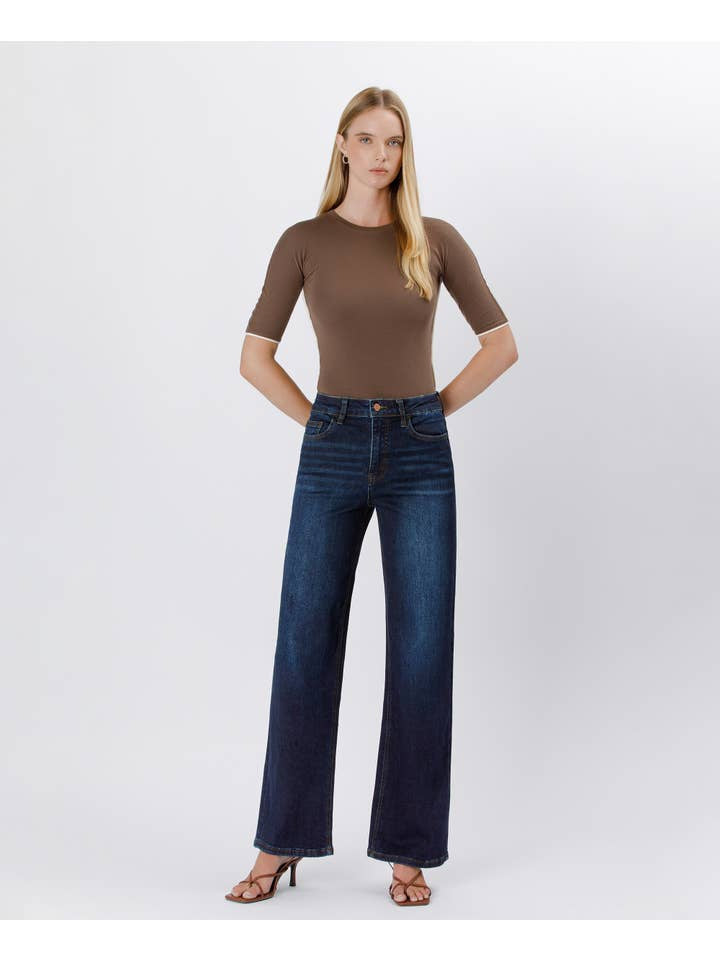 A woman stands against a plain background in a fitted brown top, heeled sandals, and Vervet's 90's Vintage Super High Rise Dark Wash Loose Jeans with wide legs, hands tucked into her pockets.