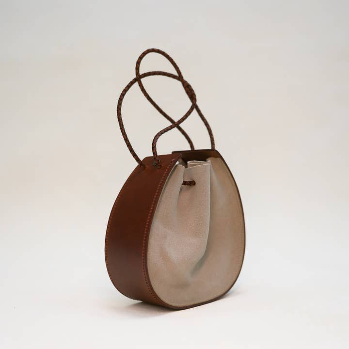 Introducing the Joi Bag by Stash in Sand Roughout—a compact, round Japanese-inspired accessory featuring a beige fabric center and brown braided handles, elegantly showcased against a simple light background.