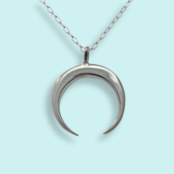 The Sterling Silver Crescent Moon Necklace by Ornamental Things features a crescent moon pendant crafted from sterling silver, hanging from a delicate, nickel-free chain and displayed against a light blue background.