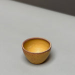 The hand-thrown Stoneware Smudge Bowl by Incausa, small and round with a yellow-brown finish, rests on a light gray surface against a blurred background.