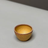 The hand-thrown Stoneware Smudge Bowl by Incausa, small and round with a yellow-brown finish, rests on a light gray surface against a blurred background.