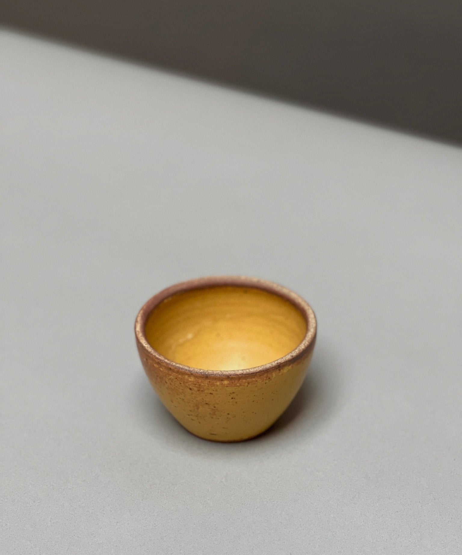 The hand-thrown Stoneware Smudge Bowl by Incausa, small and round with a yellow-brown finish, rests on a light gray surface against a blurred background.