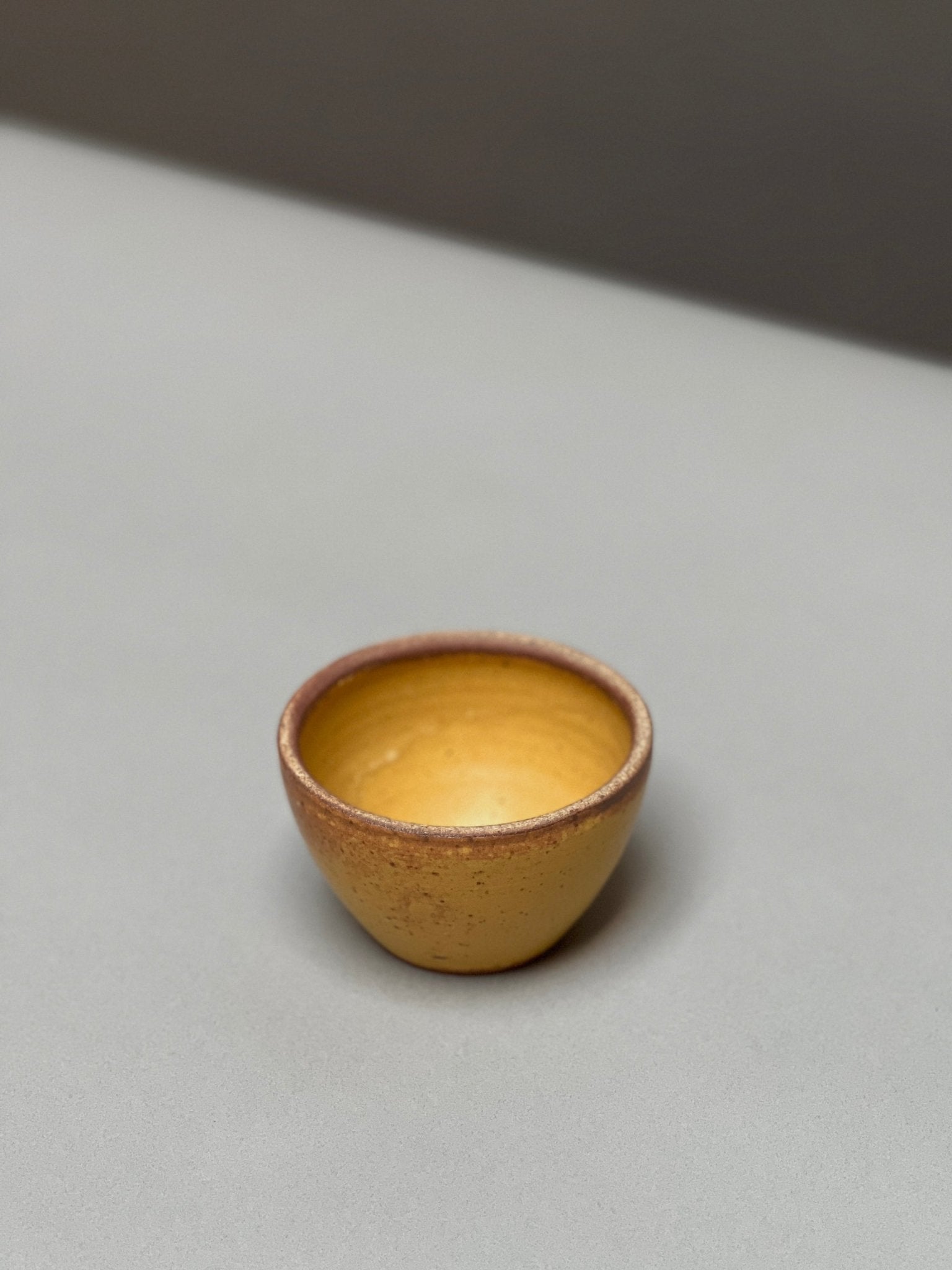 The hand-thrown Stoneware Smudge Bowl by Incausa, small and round with a yellow-brown finish, rests on a light gray surface against a blurred background.
