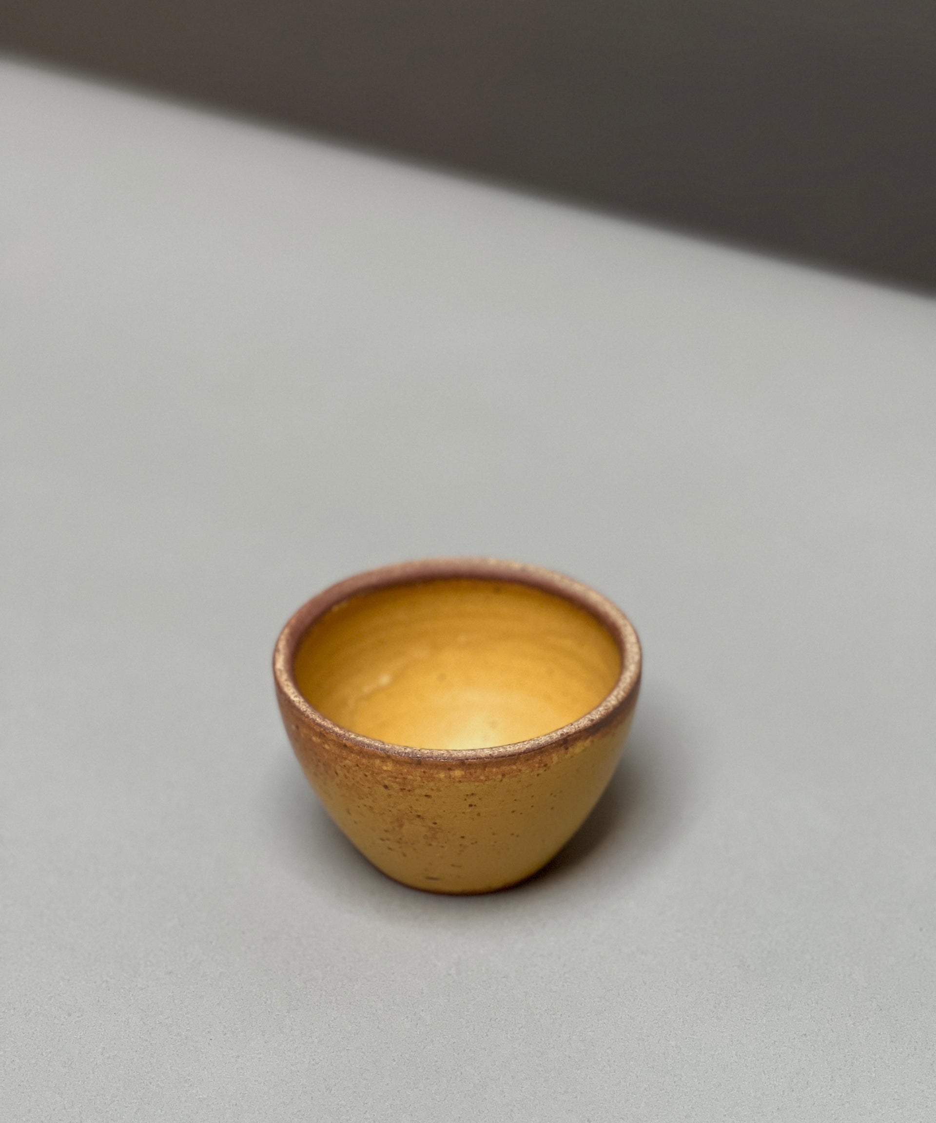 The hand-thrown Stoneware Smudge Bowl by Incausa, small and round with a yellow-brown finish, rests on a light gray surface against a blurred background.