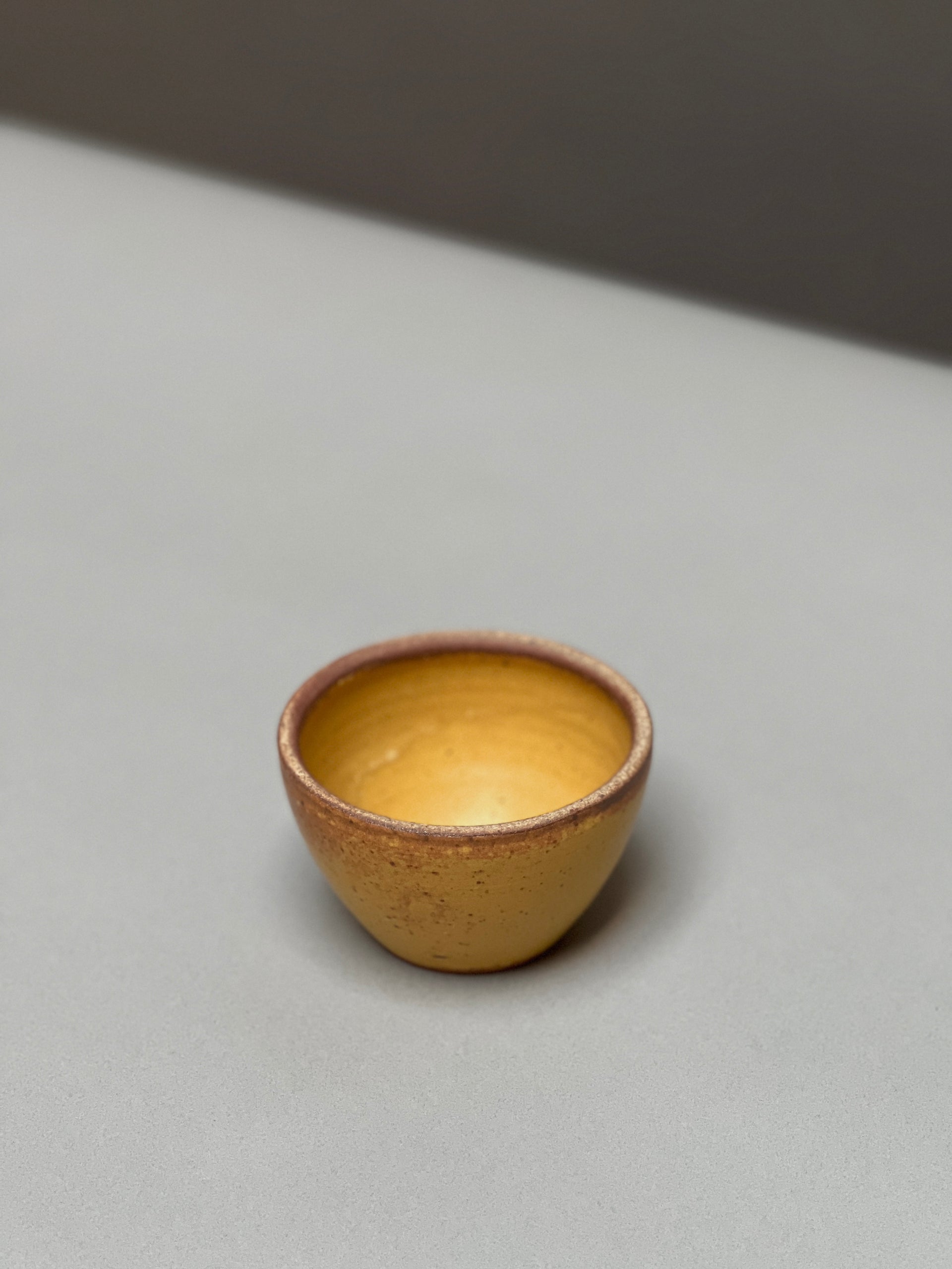 The hand-thrown Stoneware Smudge Bowl by Incausa, small and round with a yellow-brown finish, rests on a light gray surface against a blurred background.