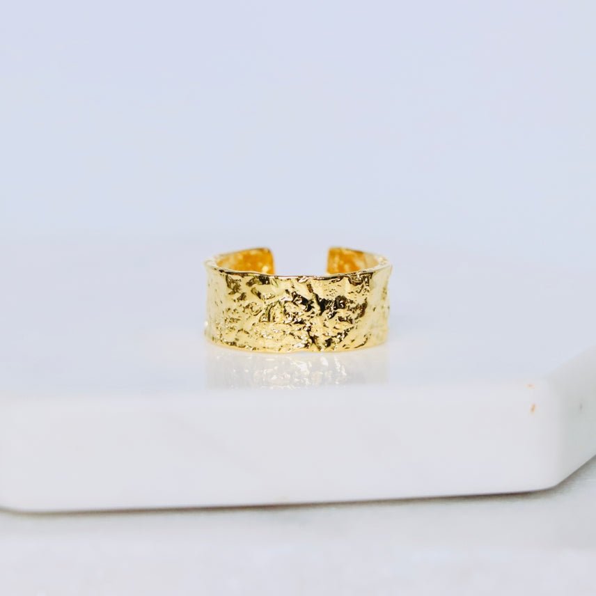 The Hammered Cuff Adjustable Ring by Mesa Blue, crafted from textured gold-plated sterling silver with an open back, rests on a white marble surface against a pale background, highlighting its elegant and versatile cuff design.