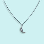 The Silver Man in the Moon Necklace by Ornamental Things features a classic crescent moon pendant with a face design, suspended from a delicate chain and showcased against a light blue background.