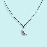 The Silver Man in the Moon Necklace by Ornamental Things features a classic crescent moon pendant with a face design, suspended from a delicate chain and showcased against a light blue background.