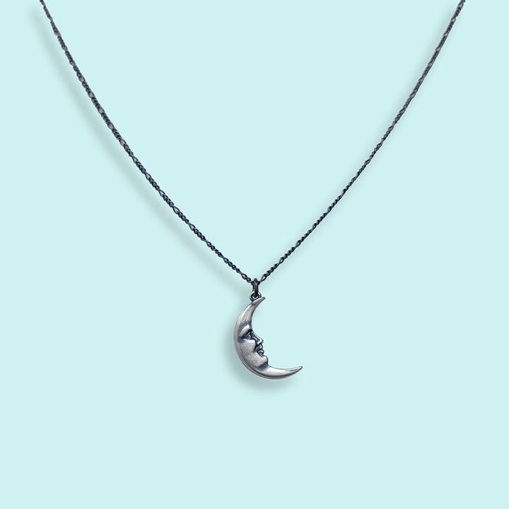 The Silver Man in the Moon Necklace by Ornamental Things features a classic crescent moon pendant with a face design, suspended from a delicate chain and showcased against a light blue background.