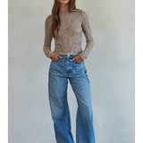 A woman stands against a plain wall wearing the By Together Knit Seamless Jacquard Mock Neck Long Sleeve Top in beige, styled with blue wide-leg jeans and black sandals, her hands resting casually in her pockets.