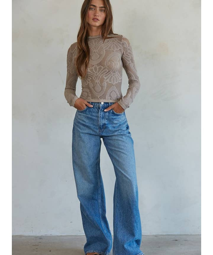 A woman stands against a plain wall wearing the By Together Knit Seamless Jacquard Mock Neck Long Sleeve Top in beige, styled with blue wide-leg jeans and black sandals, her hands resting casually in her pockets.