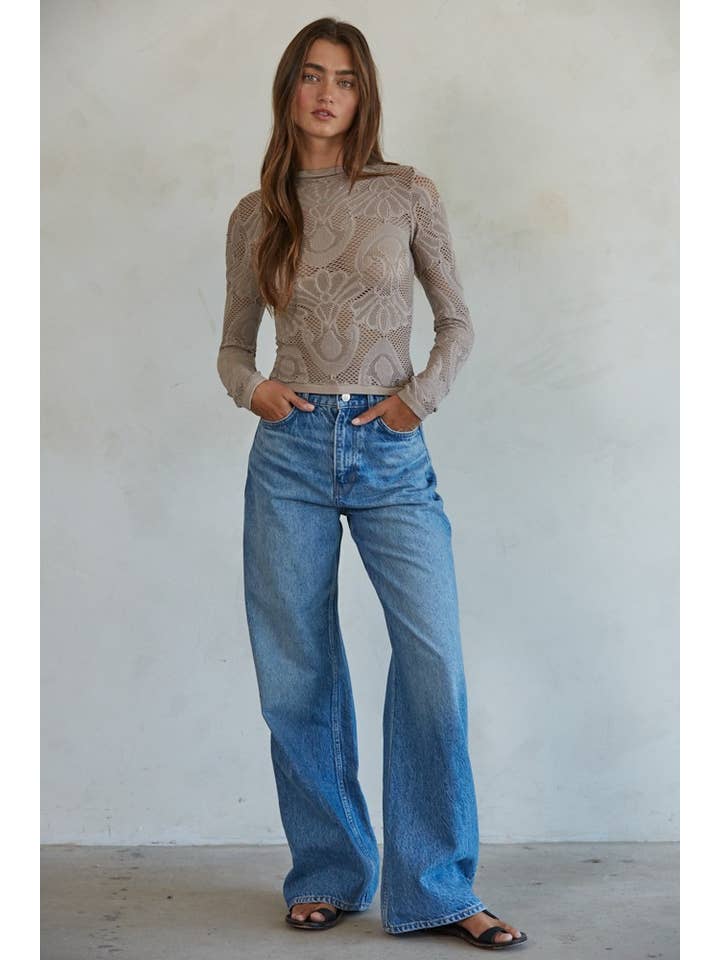 A woman stands against a plain wall wearing the By Together Knit Seamless Jacquard Mock Neck Long Sleeve Top in beige, styled with blue wide-leg jeans and black sandals, her hands resting casually in her pockets.