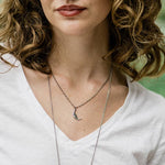 A woman with curly hair wears a white V-neck T-shirt and layered silver necklaces, including the Ornamental Things Silver Man in the Moon Necklace featuring a crescent moon pendant.
