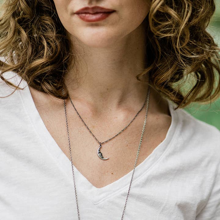 A woman with curly hair wears a white V-neck T-shirt and layered silver necklaces, including the Ornamental Things Silver Man in the Moon Necklace featuring a crescent moon pendant.