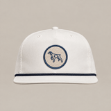The Merit Badge Hat by Ballad Originals is a white baseball cap with a beige and navy circular merit badge patch depicting a bird dog on the front, accented by a navy trim above the brim and displayed against a plain background.