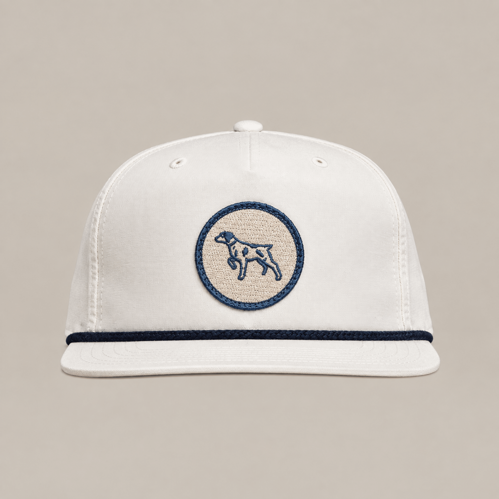 The Merit Badge Hat by Ballad Originals is a white baseball cap with a beige and navy circular merit badge patch depicting a bird dog on the front, accented by a navy trim above the brim and displayed against a plain background.