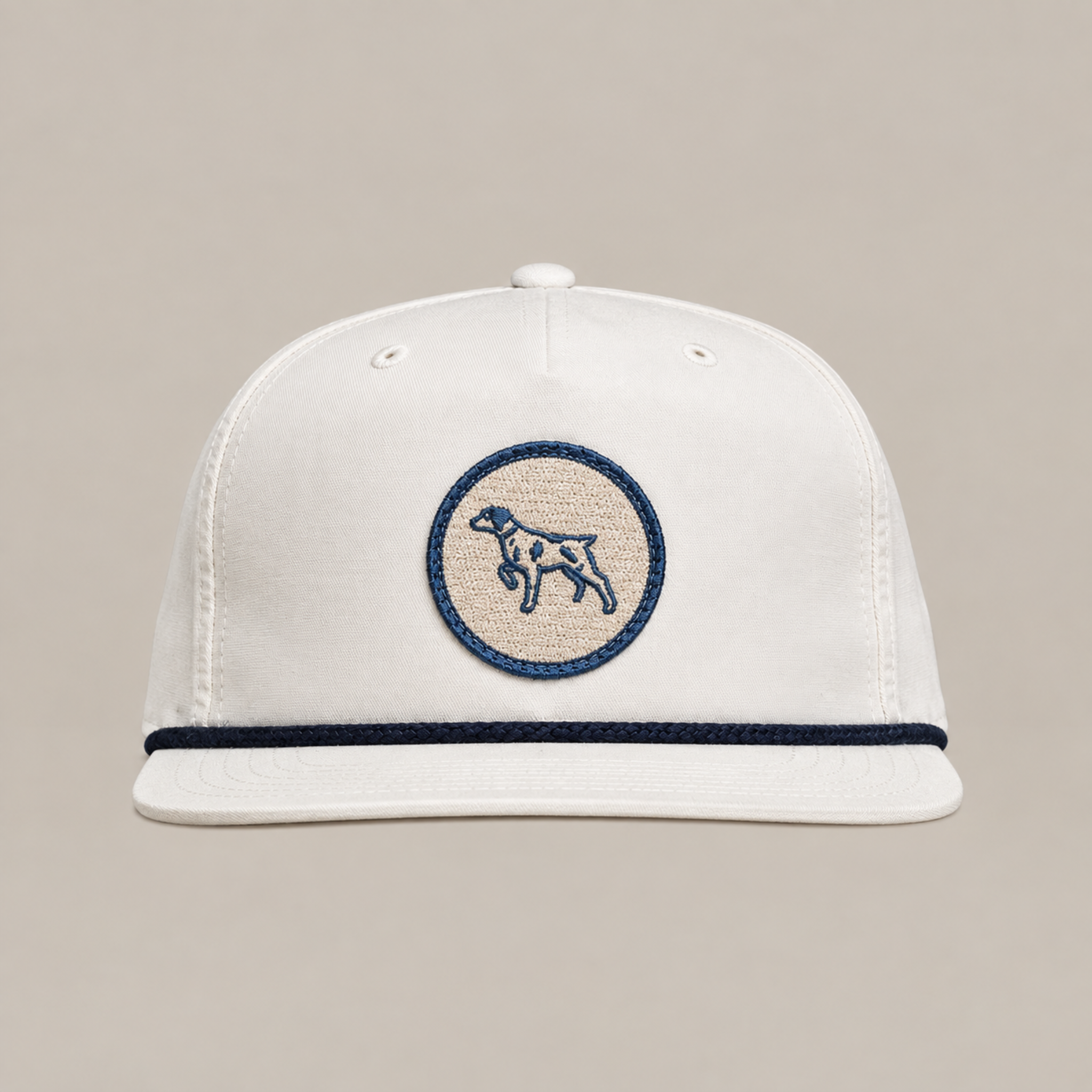 The Merit Badge Hat by Ballad Originals is a white baseball cap with a beige and navy circular merit badge patch depicting a bird dog on the front, accented by a navy trim above the brim and displayed against a plain background.