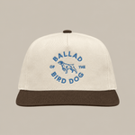The Shop Hat | Circle Clubhouse by Ballad of the Bird Dog is a beige baseball cap with a brown visor, featuring blue “Ballad of the Bird Dog” text and a central dog graphic.