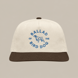 The Shop Hat | Circle Clubhouse by Ballad of the Bird Dog is a beige baseball cap with a brown visor, featuring blue “Ballad of the Bird Dog” text and a central dog graphic.
