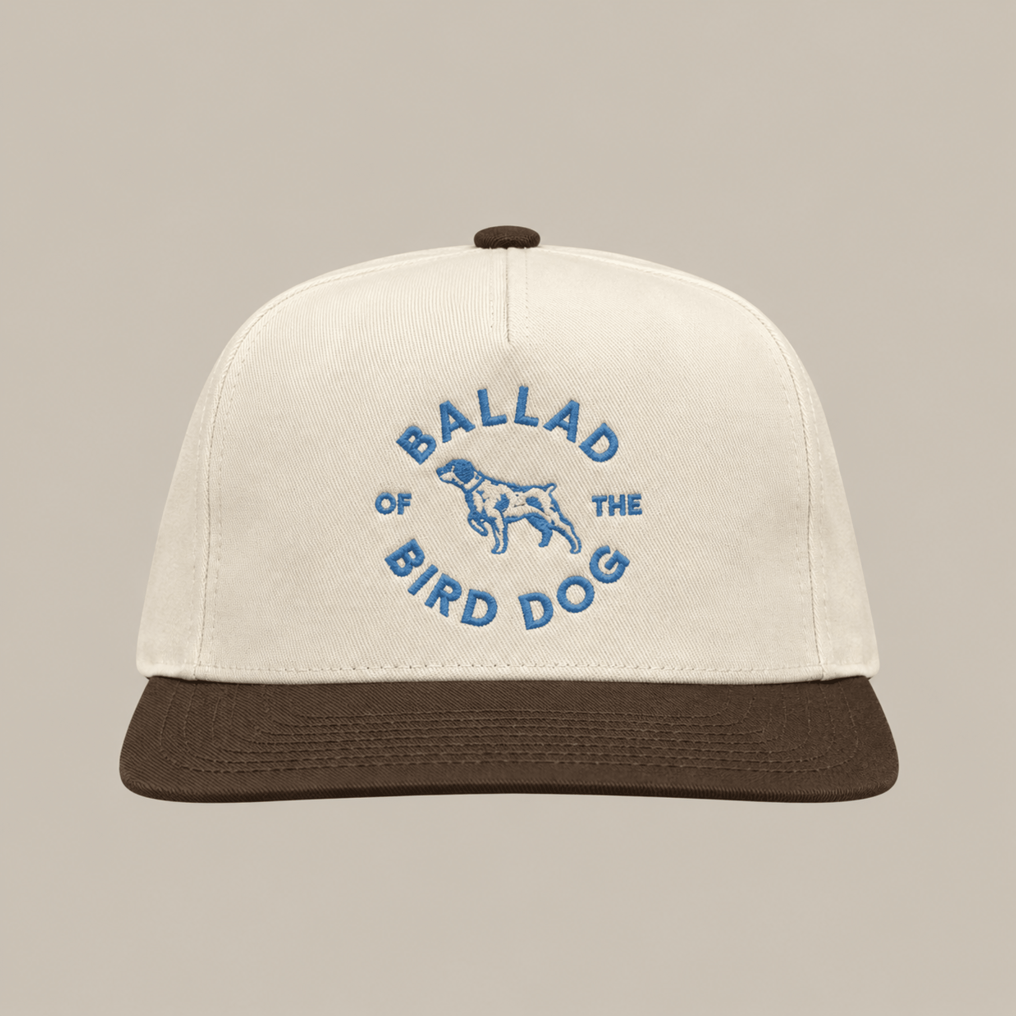 The Shop Hat | Circle Clubhouse by Ballad of the Bird Dog is a beige baseball cap with a brown visor, featuring blue “Ballad of the Bird Dog” text and a central dog graphic.