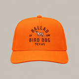 The Field Companion Hat - Ballad Originals by Ballad of the Bird Dog is a bright orange trucker hat with a mesh back, showcasing black embroidered “Ballad of the Bird Dog Texas” text and a small dog graphic on the front for a blend of classic Western style and modern comfort.