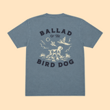 Introducing the Pheasant Flush Shirt - Ballad Originals by Ballad of the Bird Dog. This blue t-shirt features a cream and black graphic depicting a dog, birds, and plants inspired by classic upland bird hunting scenes. The phrase “Ballad of the Bird Dog” is printed on the back, making it a standout choice for any pheasant hunting shirt collection.