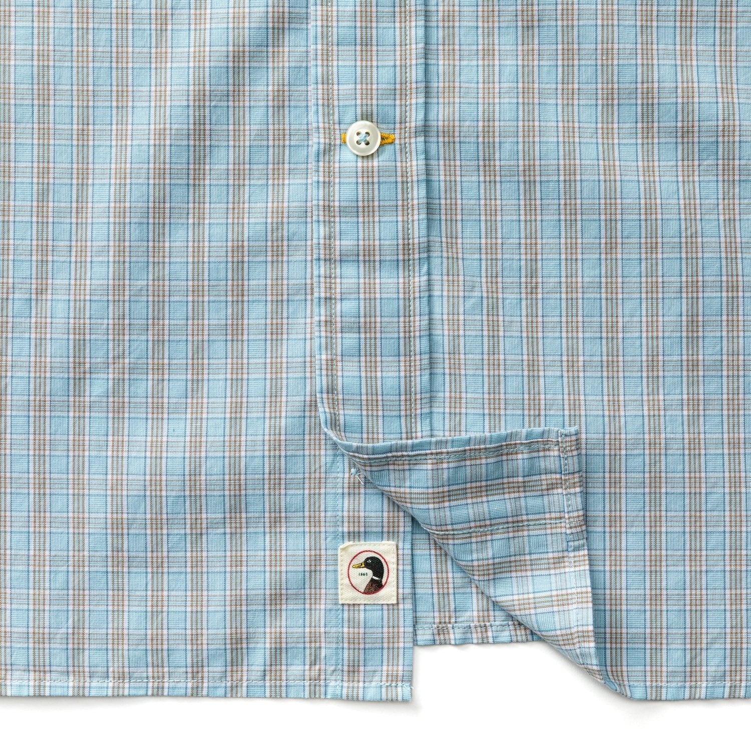 A close-up of the Duck Head Cotton Cashmere Poplin Sport Shirt in blue plaid with tan and white lines, featuring a white button and a small toucan logo tag near the bottom hem.