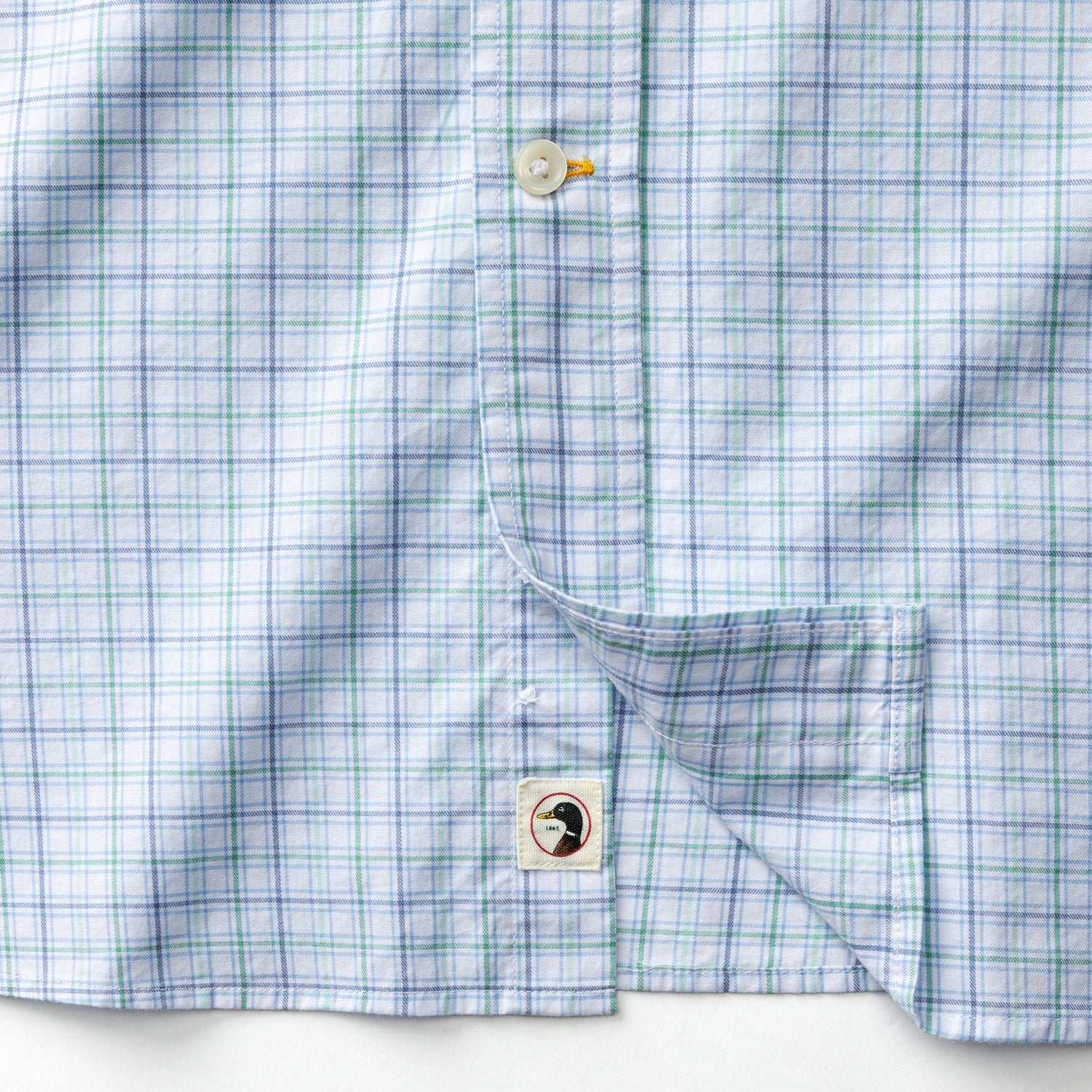 A close-up of the Cotton Twill Sport Shirt by Duck Head in light blue and green plaid, featuring buttons, a pocket, and a small square tag with a black duck graphic near the bottom hem.