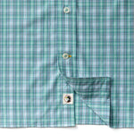Close-up of the Duck Head Performance Poplin Sport Shirt in green and blue plaid, with one side lifted to reveal a small tag displaying the black and white Duck Head logo.
