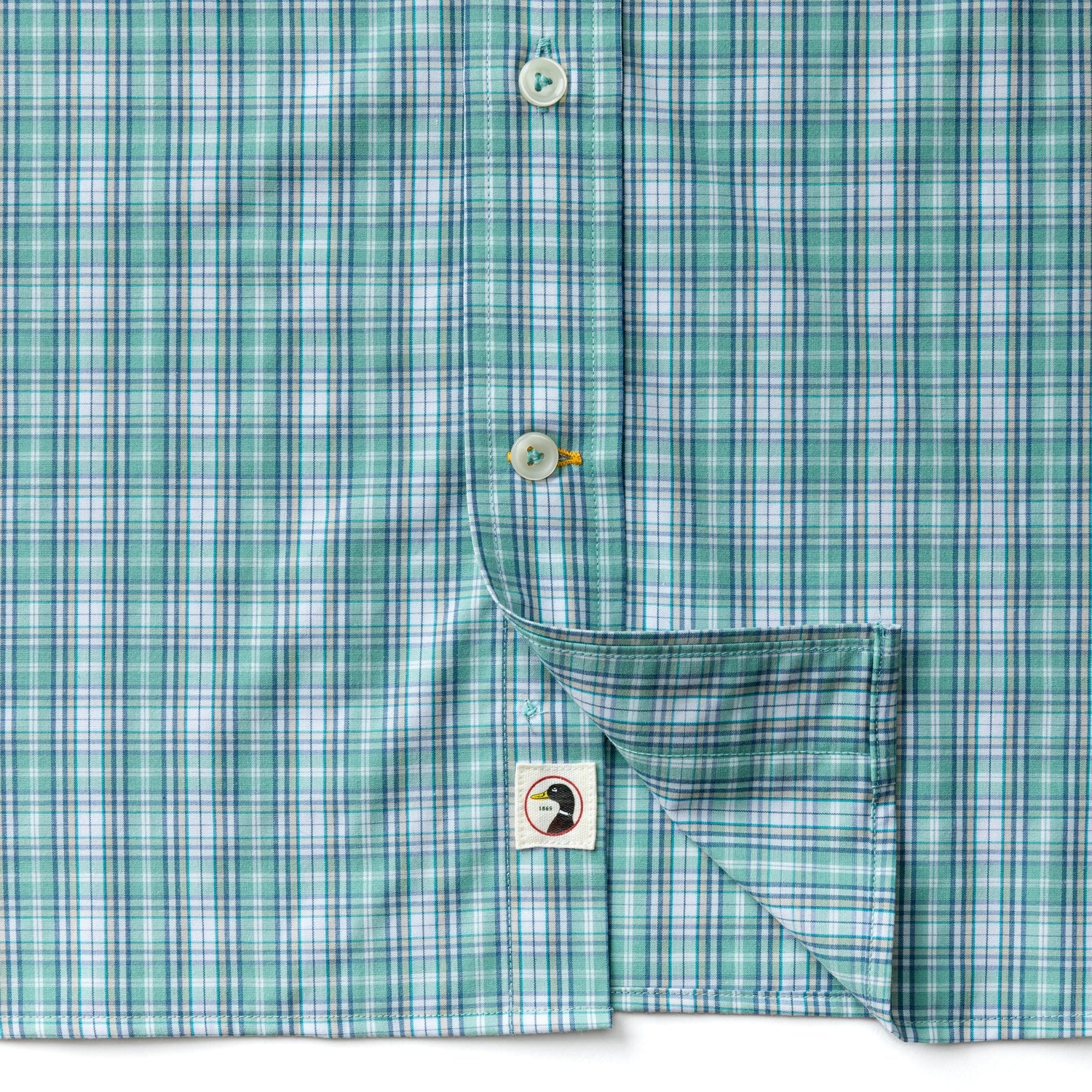 Close-up of the Duck Head Performance Poplin Sport Shirt in green and blue plaid, with one side lifted to reveal a small tag displaying the black and white Duck Head logo.