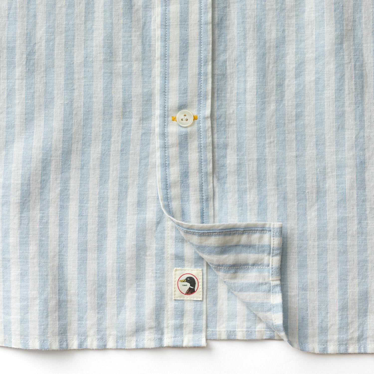 Close-up of the Duck Head Linen Cotton Oxford Sport Shirt, featuring blue and white vertical stripes, a button-down collar, and a small bird logo patch near the hem.