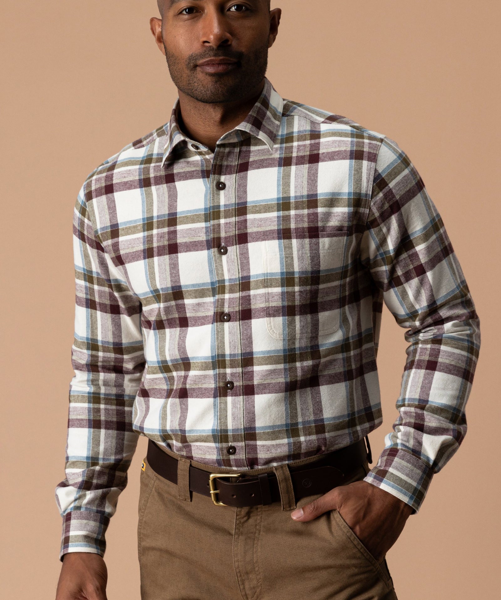A man wears a Huckleberry Cotton Flannel Sport Shirt by Duck Head in white, maroon, and blue plaid, tucked into brown trousers with a brown leather belt.