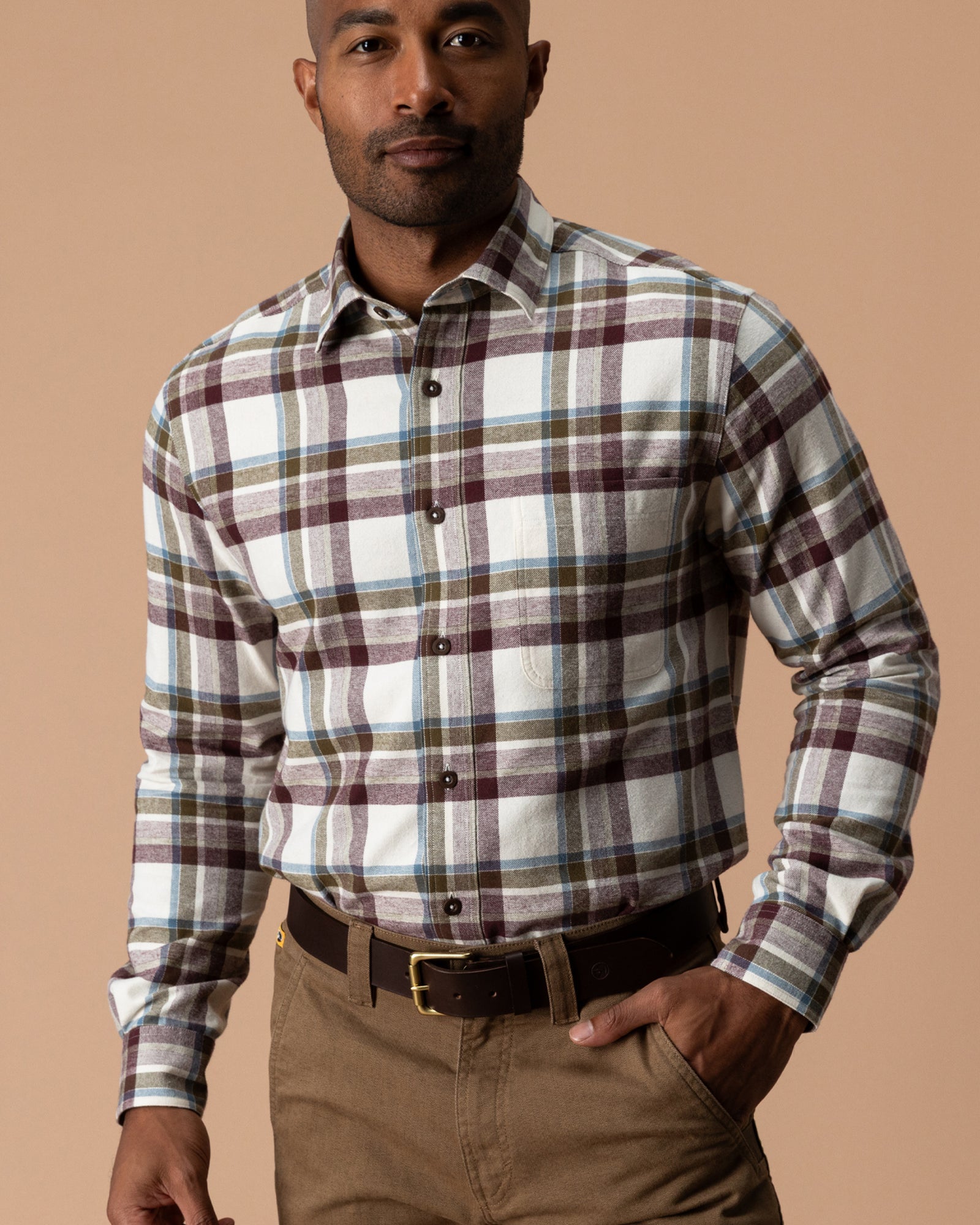 A man wears a Huckleberry Cotton Flannel Sport Shirt by Duck Head in white, maroon, and blue plaid, tucked into brown trousers with a brown leather belt.