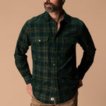 A man wears a Duck Head Corduroy Snap Shirt in a dark green and navy plaid cotton corduroy with two chest pockets.