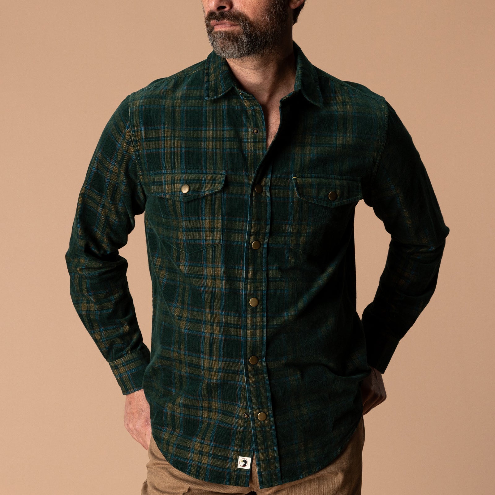 A man wears a Duck Head Corduroy Snap Shirt in a dark green and navy plaid cotton corduroy with two chest pockets.