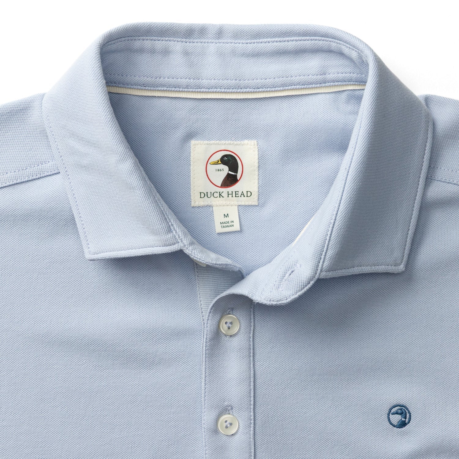 The Duck Head Hanover Pima Cotton Pique Polo features a light blue hue, classic collar, button placket, small chest logo embroidery, and a branded label inside the collar.