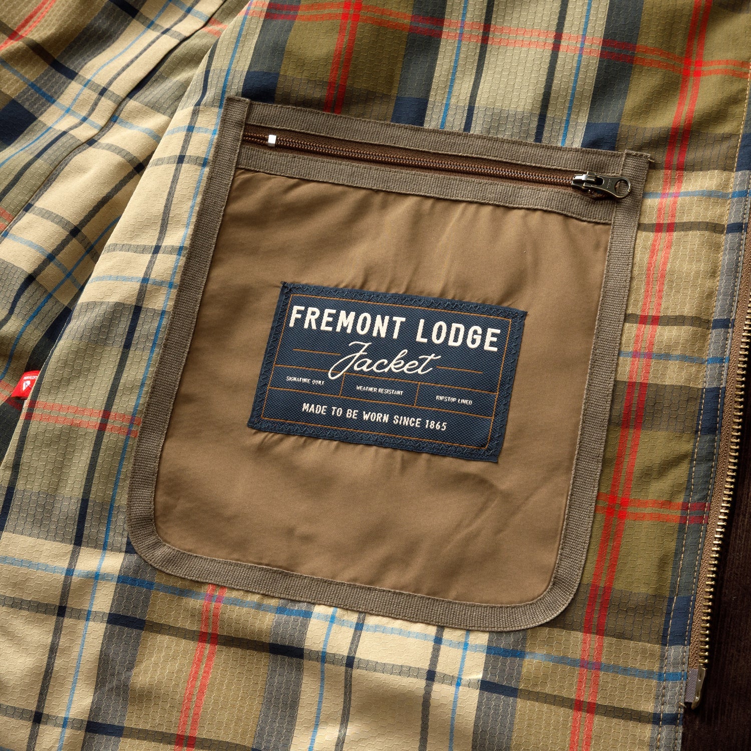 Interior view of a Duck Head Fremont Lodge Performance Quilted Jacket with a plaid lining and a zippered pocket featuring a label that reads “Fremont Lodge Jacket, made to be worn since 1865.”.