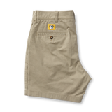 A pair of folded Duck Head 7in Gold School men’s chino shorts with a buttoned back pocket and a yellow globe logo patch above the pocket.