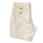 A pair of Duck Head 7in Gold School men's chino shorts in beige is folded to show the back with a single buttoned pocket and a yellow patch above it. The shorts feature a classic 7-inch inseam.