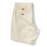 A pair of Duck Head 7in Gold School men's chino shorts in beige is folded to show the back with a single buttoned pocket and a yellow patch above it. The shorts feature a classic 7-inch inseam.