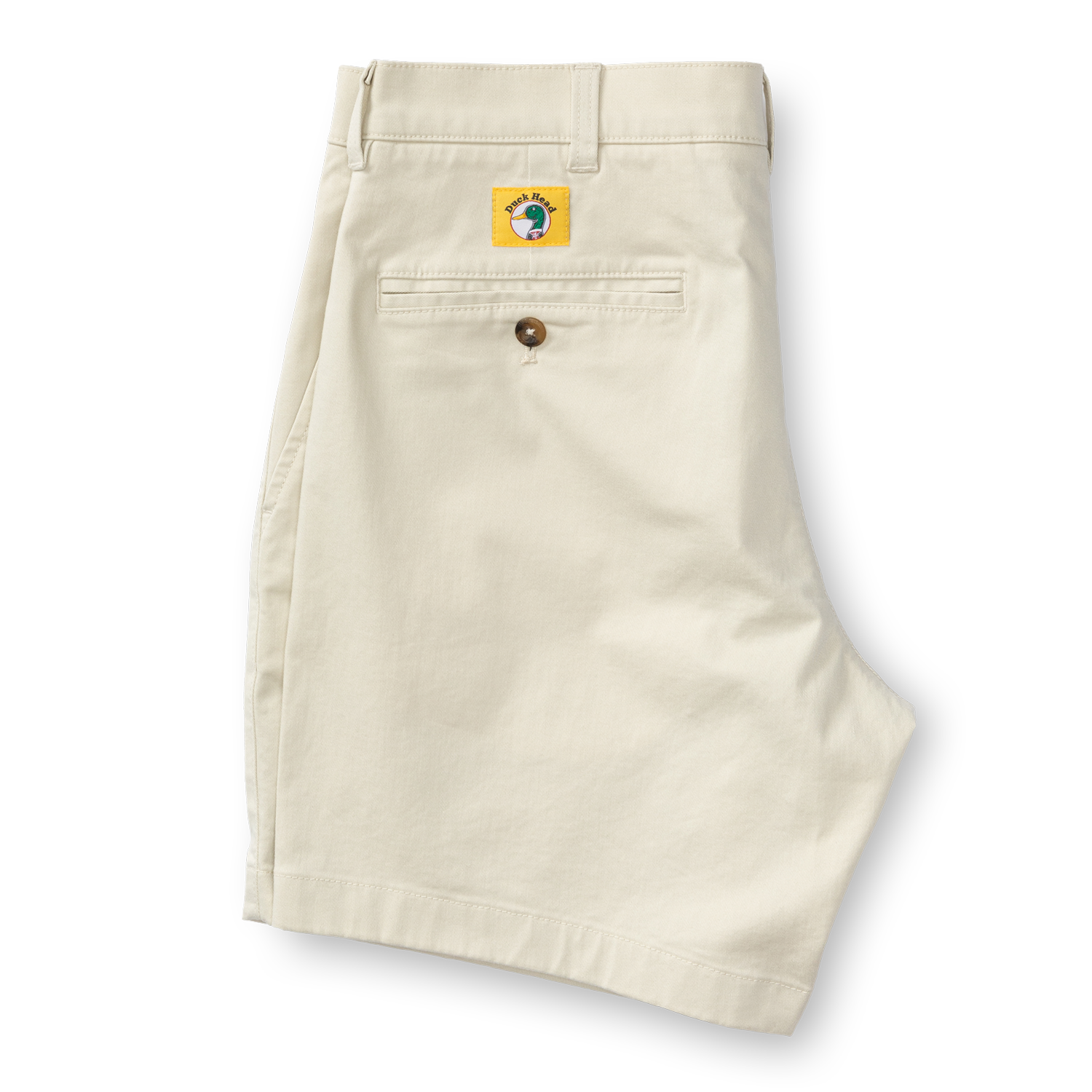 A pair of Duck Head 7in Gold School men's chino shorts in beige is folded to show the back with a single buttoned pocket and a yellow patch above it. The shorts feature a classic 7-inch inseam.